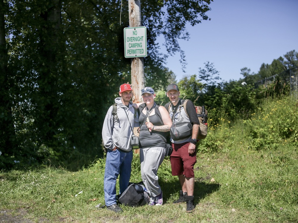 Three people stand in a grassy area in front of a telephone or hydro pole. On the pole, there is a sign that reads, “No Overnight Camping Permitted.”