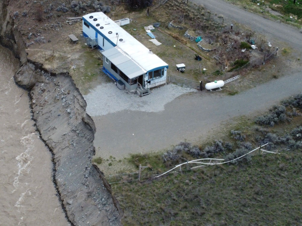 An aerial photo of the property currently threatened by the river. The mobile home’s metal roof shines in the light. Five metres from the home’s front door the land cuts away to a sandy embankment which leads to the muddy waters of the Nicola River. The property’s fence line juts out over the edge.