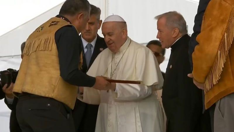 The Pope is facing Chief Terry Teegee. Chief Teegee, whose back is towards the camera, gives the pope a long wooden stick.