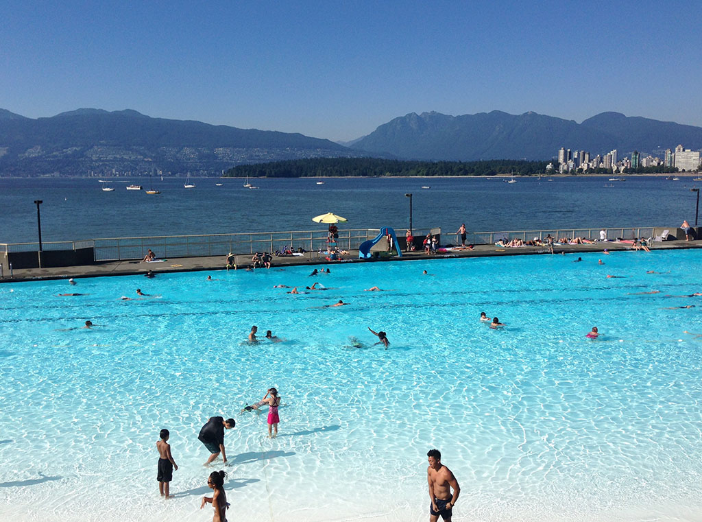 Kits Pool, a large, extremely pale blue basin of water busy with people swimming and wading, serves as foreground to the dark blue waters of English Bay and rising mountains on a clear day.