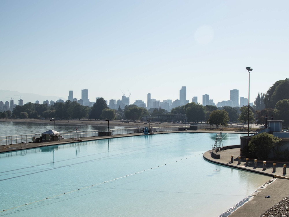 Kits Pool, a large, extremely pale blue basin of water empty of people, serves as foreground to the dark blue waters of English Bay and rising mountains on a clear day.