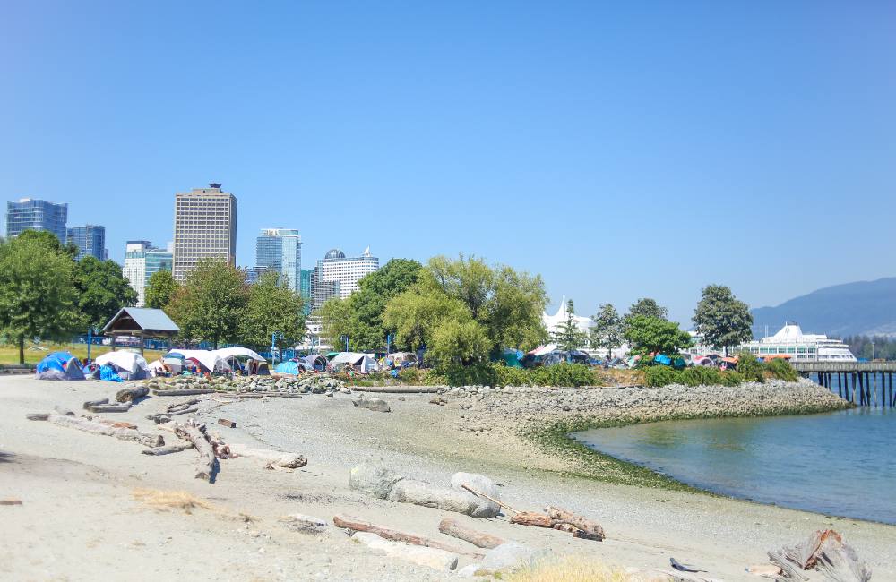 A beach is in the foreground and the CRAB Park tent city can be seen against a sparse treeline. Downtown Vancouver is in the background. The sky and water are blue.