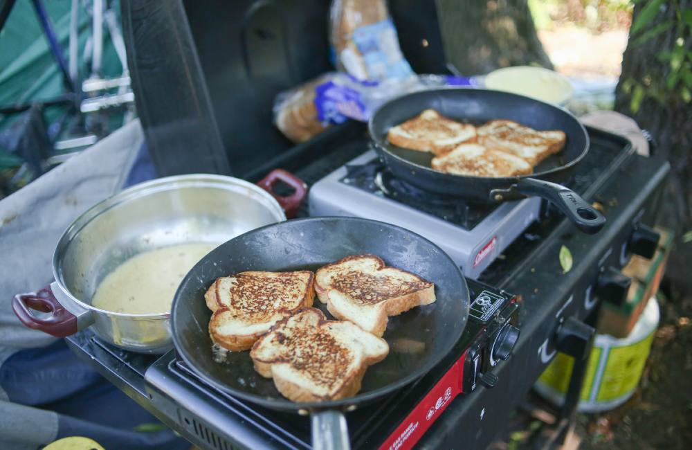 Two pans with three slices of French toast each are cooking on outdoor stoves. Behind them is a large bowl with egg batter.