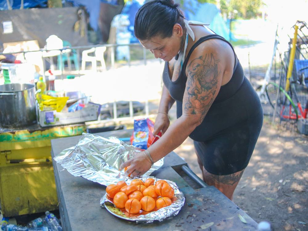 A woman with a red ceramic knife halves oranges and adds them to a plate. She’s standing in an outdoor kitchen in a park under the shade of a tree.