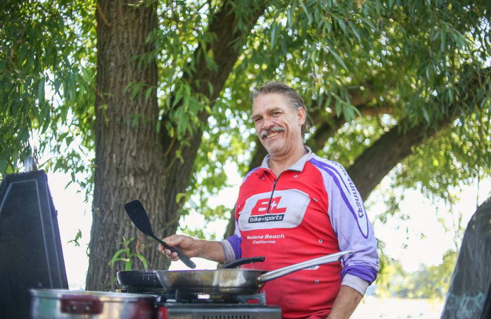 A man in a bike shirt with greying hair and mustache has a big smile on his face. He’s holding a spatula in front of an outdoor stove under the shade of a tree.