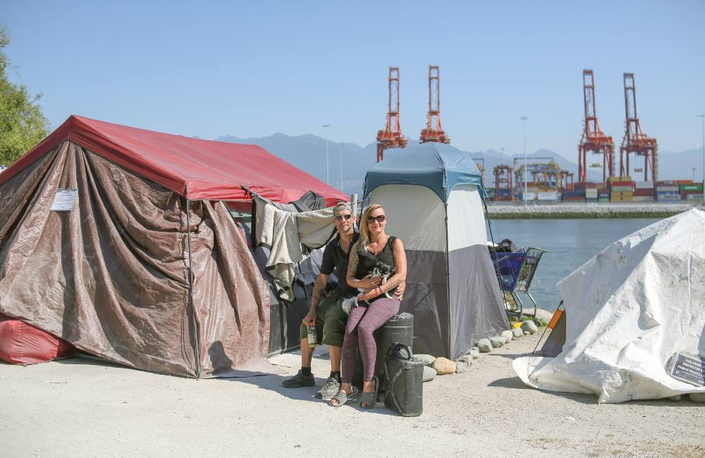 A couple holding a dog sits on large, black containers of water beside their tent residence on the beach, which consists of one large residential tent and a smaller shower tent. Behind them is the waterfront, with tall red cranes at the nearby port and a backdrop of mountains.
