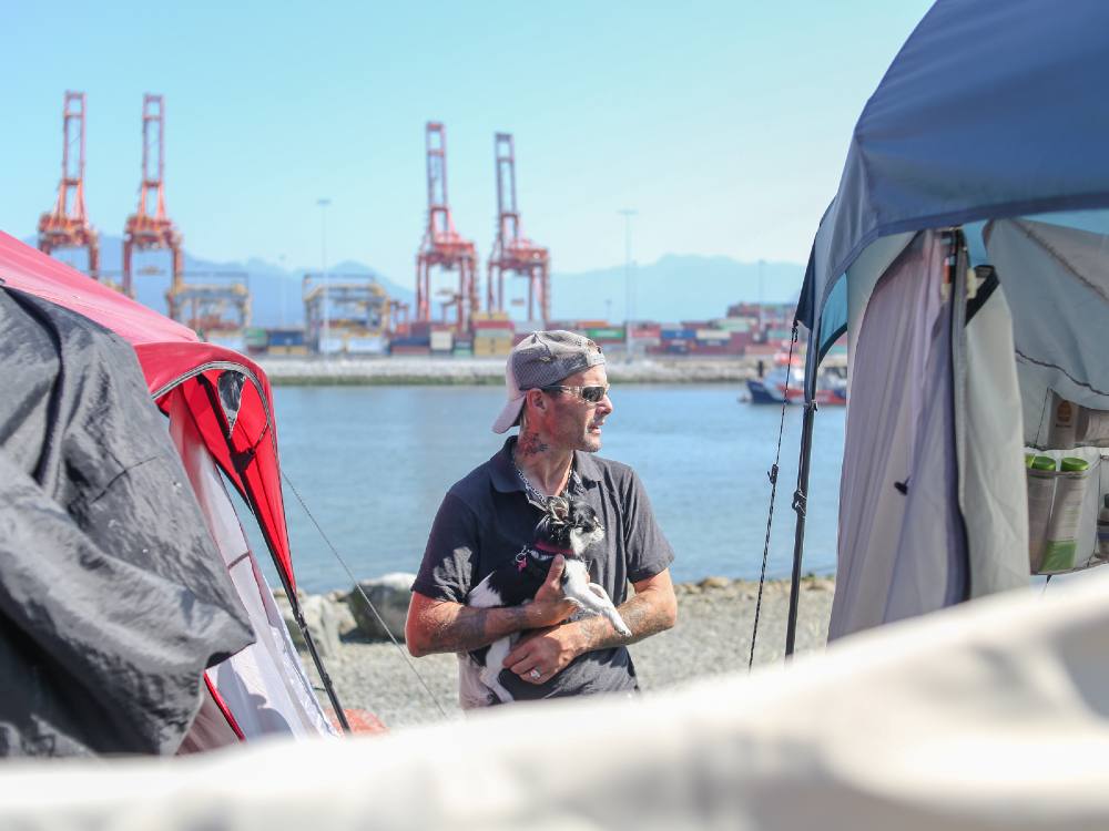 A man wearing sunglasses and a cap is standing between two tents with a dog in his arms. Behind him is the water of the inlet, the red cranes of a nearby and Vancouver’s North Shore mountains.