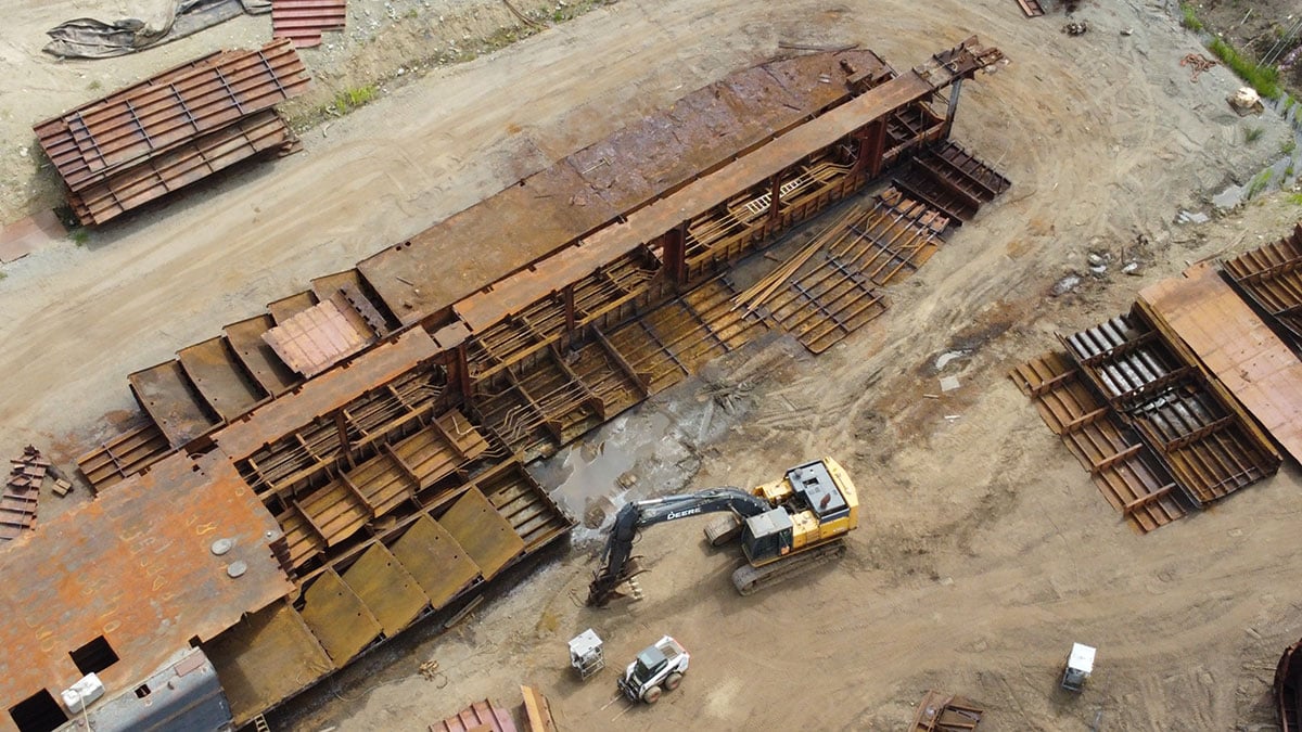 A yellow excavator sites in front of the remains of a partially dismantled rusty barge, sitting on sandy, muddy ground.