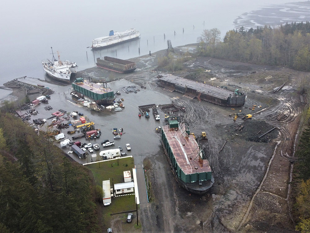 An aerial shot shows an oceanside site with several ships and barges in various stages of being dismantled. There is a paved parking lot, but three large vessels sit on muddy land.