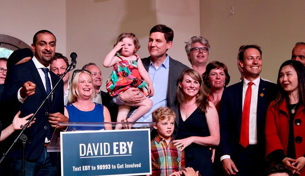A beaming David Eby stands on a stage holding a child and surrounded by cheering people, with a sign telling people how to contact his campaign.
