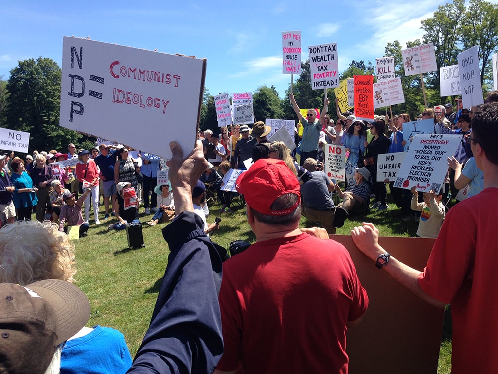 A group of mainly white, older people gather in a park setting holding signs saying, among other things, ‘NDP = Communist Ideology’ and ‘Don’t Tax Wealth, Tax Poverty Instead’