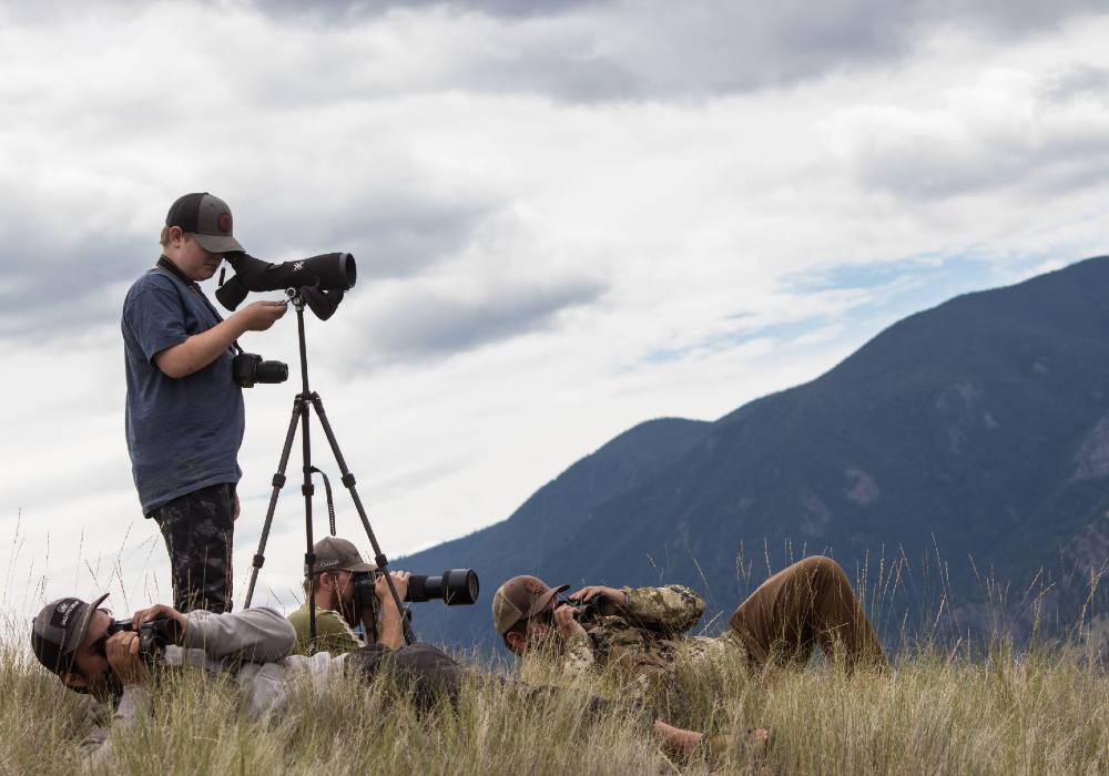 Three men lie in high grass peering through binoculars while another stands behind a telescope on a tripod.