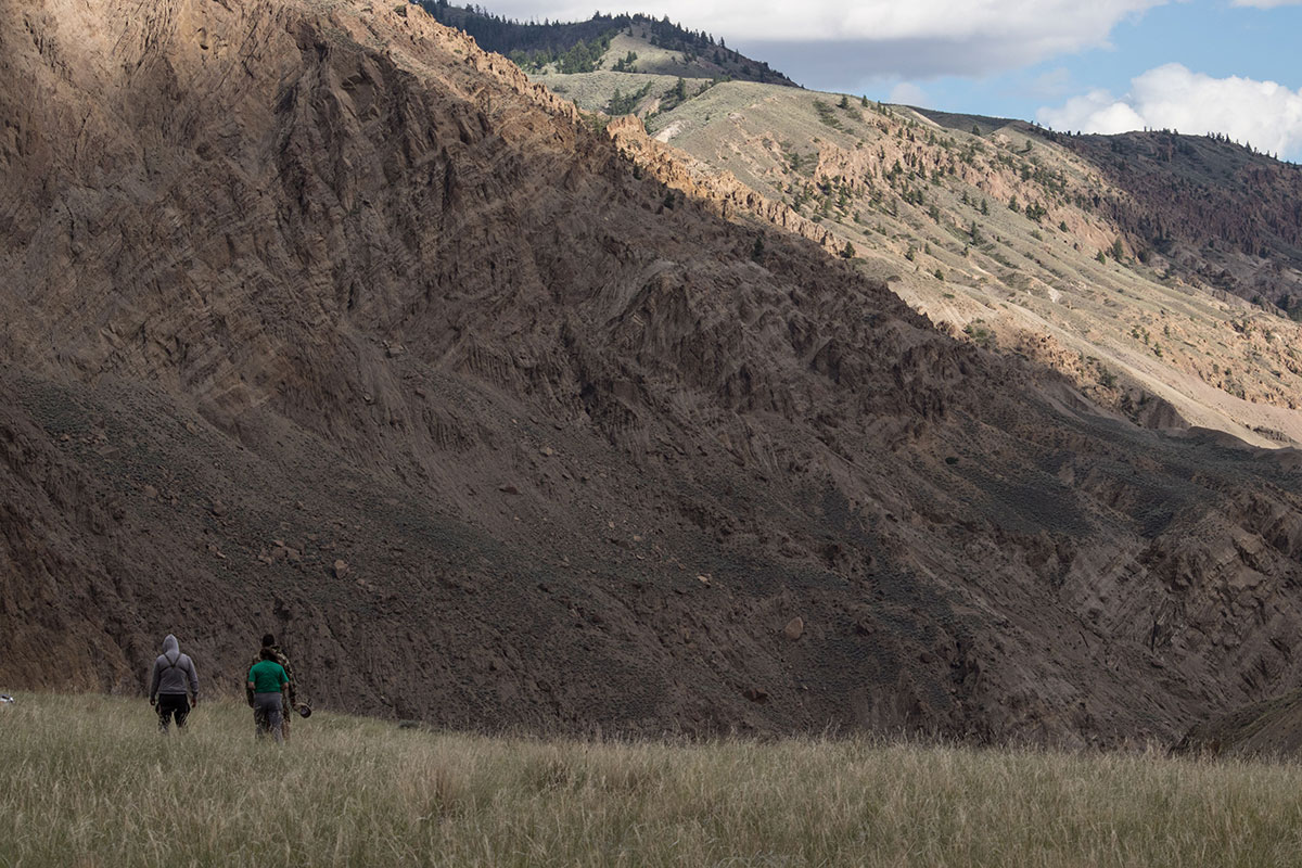 Three people walk on a grassy landscape flanked by mountains. 