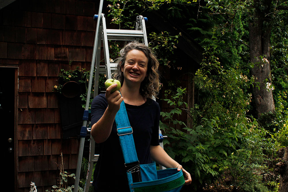 Michelle Gamage stands in front of a ladder, wearing an apple-picking bag and holding a green apple out towards the camera. 