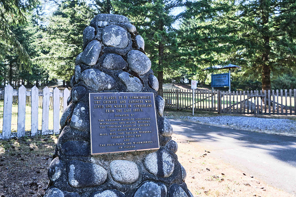 A memorial monument made of stone is mounted with a plaque that reads “This plaque is to commemorate the Chinese and Japanese who lived and worked in Cumberland from the late 1800s.” The entrance for the Chinese cemetery is in the background to the right.