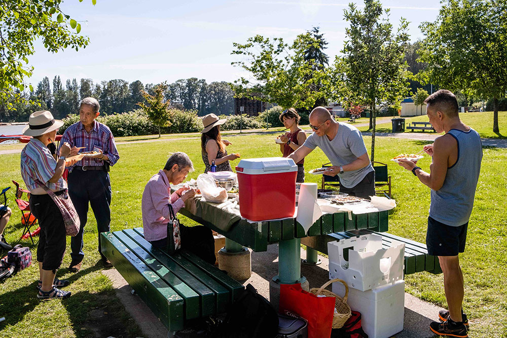 A group of people gather around a picnic table at Riverfront Park in East Vancouver on a sunny day.