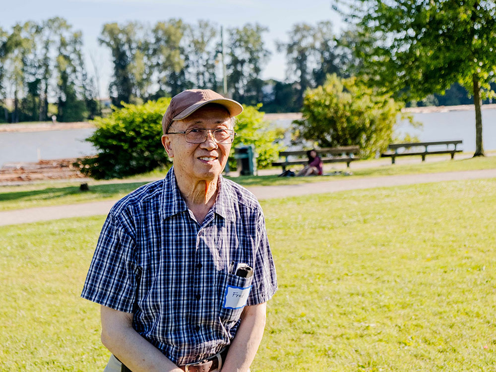 Fred Leung is wearing a tan ball cap, glasses, a blue plaid shirt and khakis. He is smiling at the camera. In the background is green grass, picnic tables and a river. 