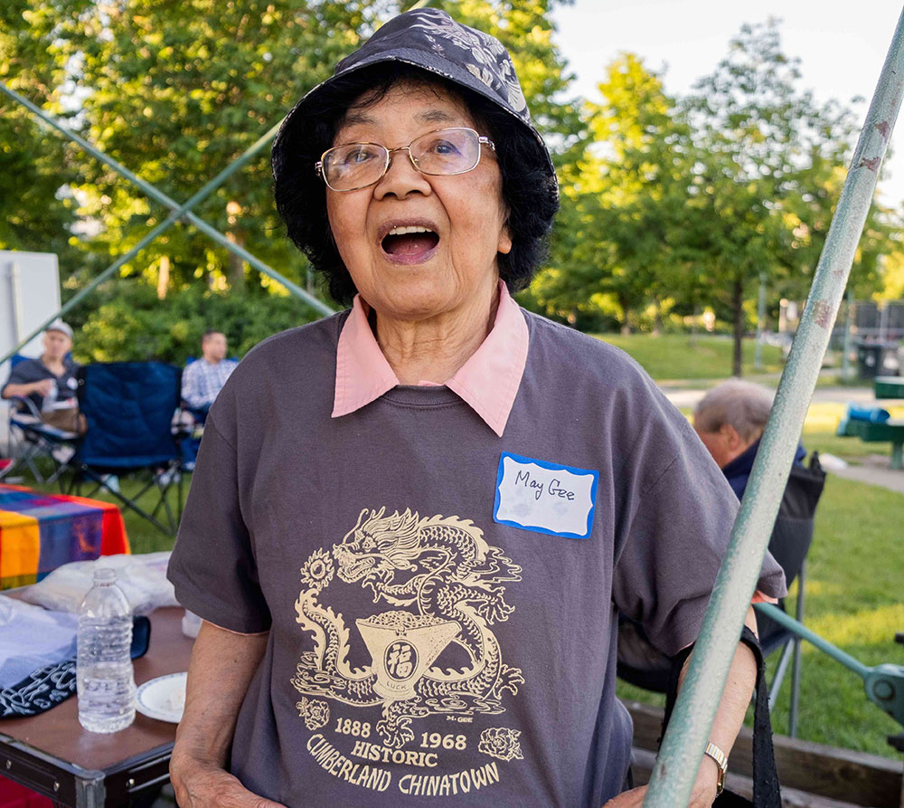 May Gee is wearing a bucket hat and smiling. She is showing off a dusty blue shirt emblazoned with a gold dragon logo that reads “Cumberland historic Chinatown, 1888-1968.”