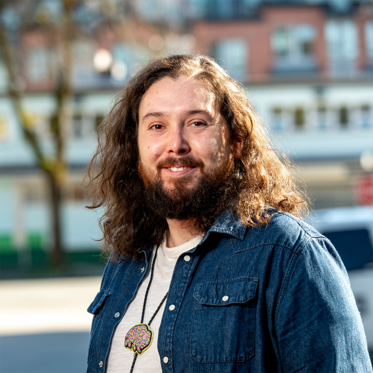 Matthew Norris is wearing a denim shirt over a white t-shirt. He is standing against a blurred urban background.