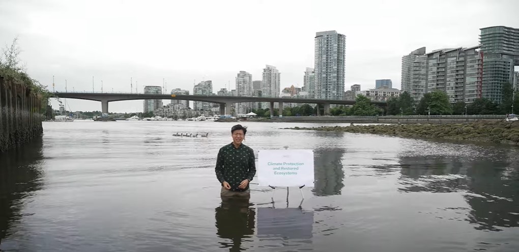 Uytae Lee stands waist-deep in Vancouver’s False Creek near Habitat Island in the Olympic Village. The sky and water are grey. The Cambie Bridge is in the background.