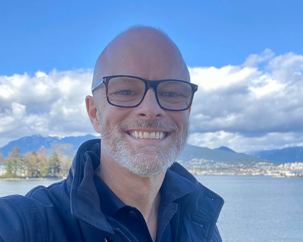 Mark Marissen is wearing glasses and a blue jacket. He is standing near the water. The sky and water are blue.