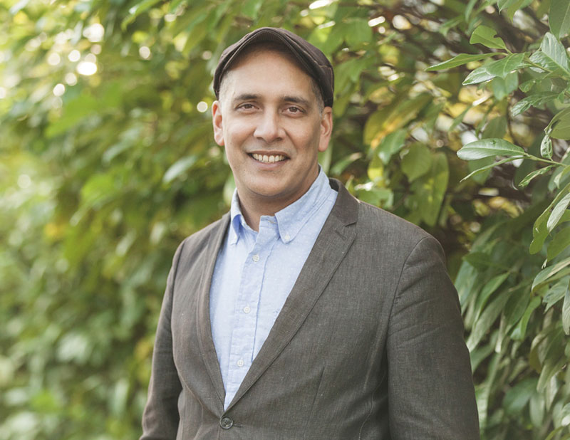 Pete Fry is wearing a twill blazer, blue shirt and cap. He is standing against a lush green background of rhododendron plants. 