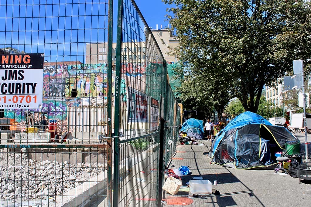 A wire fence blocks access to a rubble-filled construction site. Two large tents are on the adjacent sidewalk.
