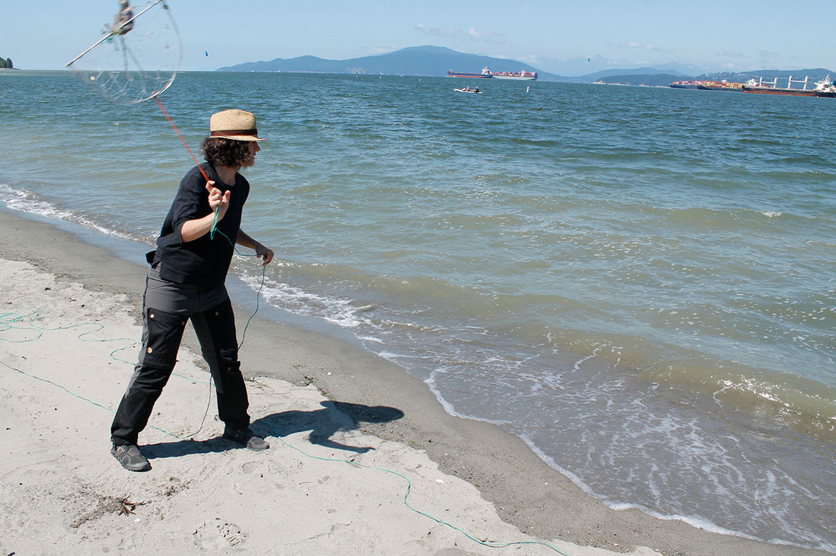 A woman in a straw hat swings a half-moon crab trap around on a red rope, preparing to throw it into the calm waters off a sandy beach. 