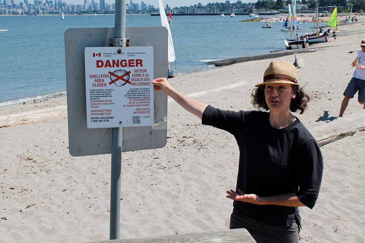 A woman wearing a straw hat points to a sign on a beach that warns against eating shellfish in the area. The Vancouver skyline is visible in the background. 