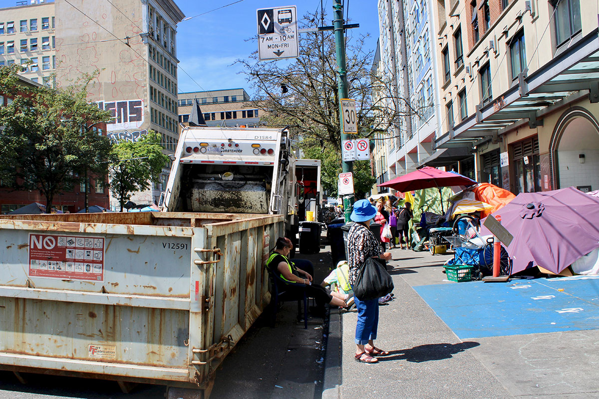Tents, umbrellas and people’s belongings line the sidewalk on East Hastings Street in front of buildings. City workers in reflective vests squat in front of a dumpster and a garbage truck.