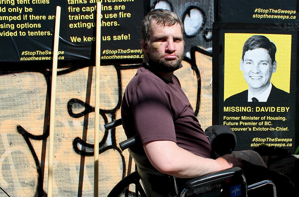 A man with brown hair and a beard, wearing a purple shirt and sitting in a wheelchair, looks into the camera. Behind him are signs calling for an end to street sweeps and criticizing NDP leadership candidate David Eby.