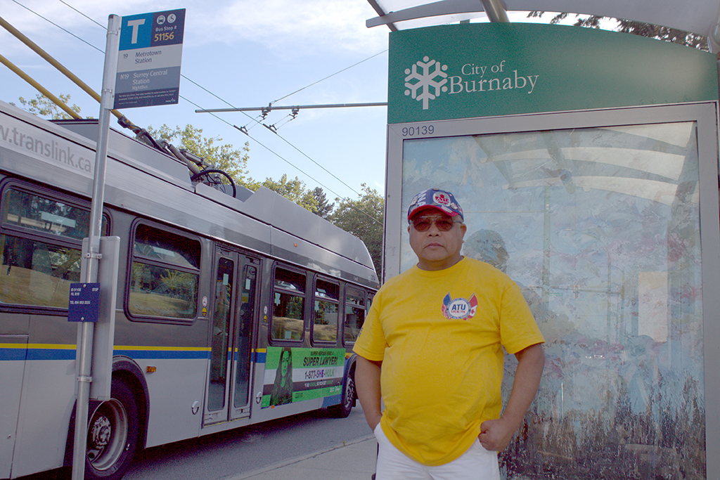 Arturo Buban stands at a bus stop in Burnaby. He is wearing a yellow ATU union T-shirt and a red, white and blue baseball cap from the same union. He is standing near a bus stop for the 19 Metrotown Station bus, which will take him to work on the North Shore.