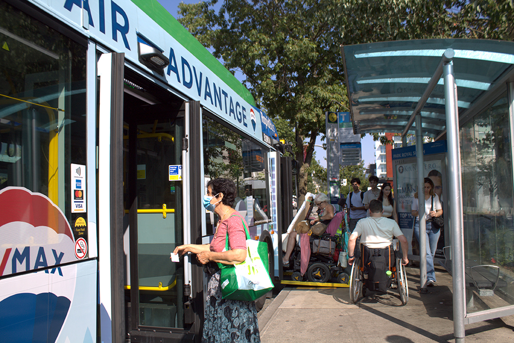 A busy, sunny bus stop in West Vancouver is crowded with passengers as they board the R2 rapid bus. A woman with a tote bag is in the foreground, boarding at a side door. A person with a wheelchair approaches the front door. The bus is covered with an advertisement for Remax realtors.