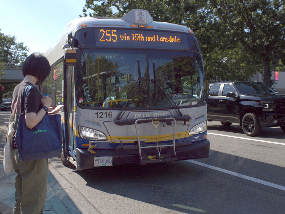 A bus with a sign that reads “255 via 15th and Lonsdale” slows for a passenger waiting to board to the left of the frame. The bus is blue and grey with a yellow digital sign. The waiting passenger has short black hair and is carrying a blue tote bag.