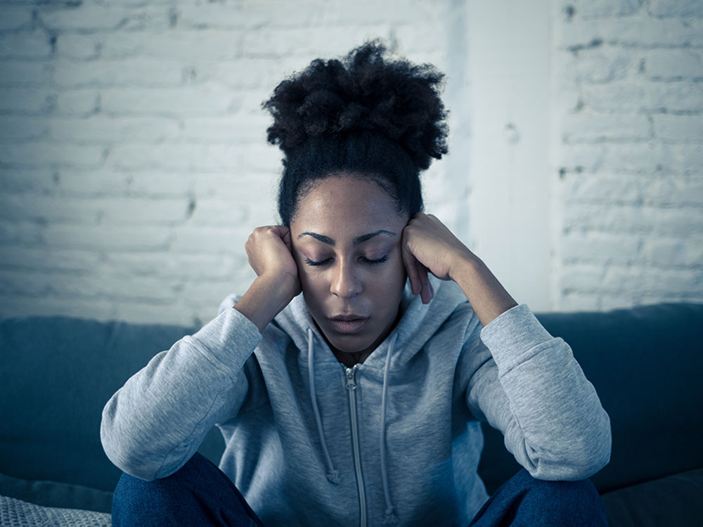 A Black woman sits on a couch with her eyes closed, and her hands against her cheeks. She looks depressed.