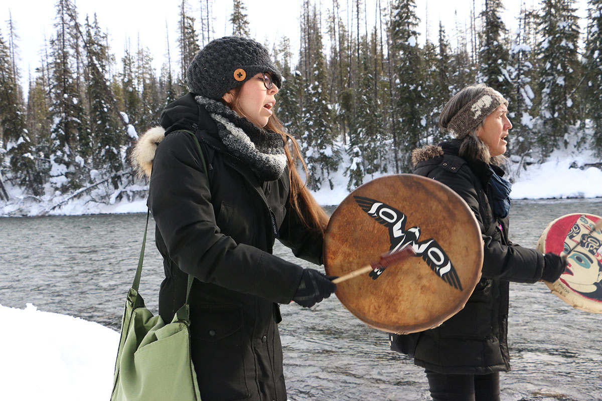 Two women drum in winter clothing drum next to a fast-moving, slate-blue river. There is snow on the ground. 