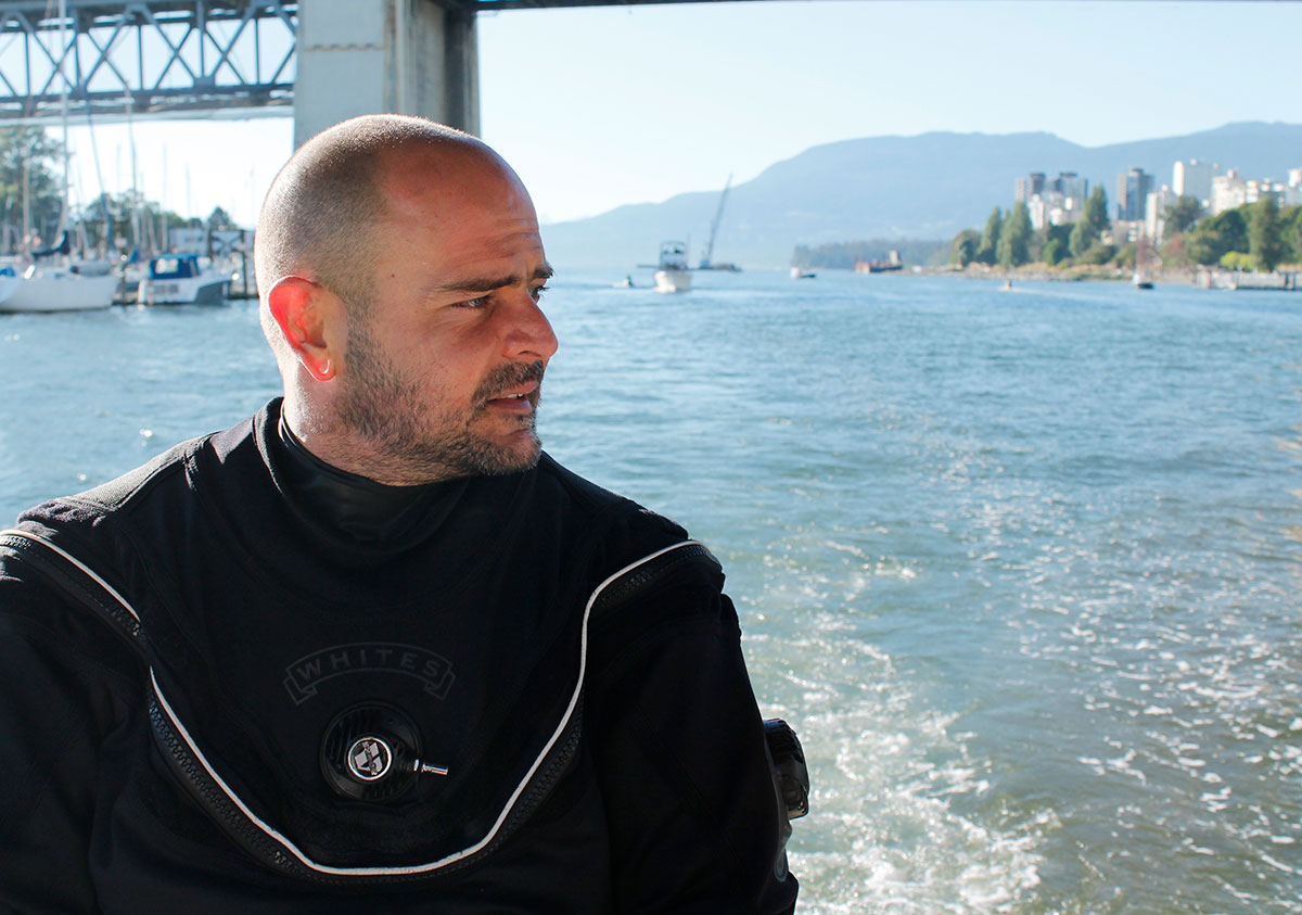A man in a black drysuit sits on the edge of a boat with the Burrard Street bridge behind him.