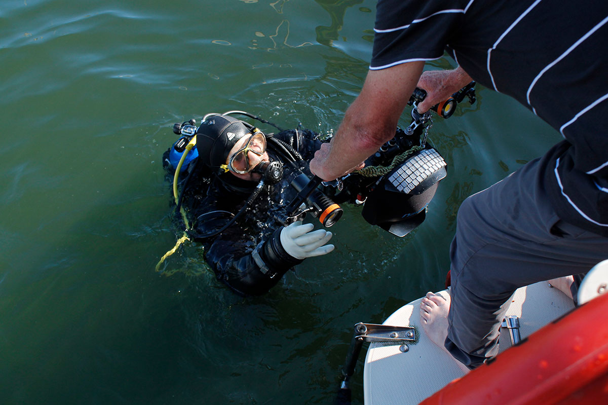 A man leans over the water to hand a large underwater camera to a diver in the water. The diver is reaching their white gloved hands up to receive the camera. 