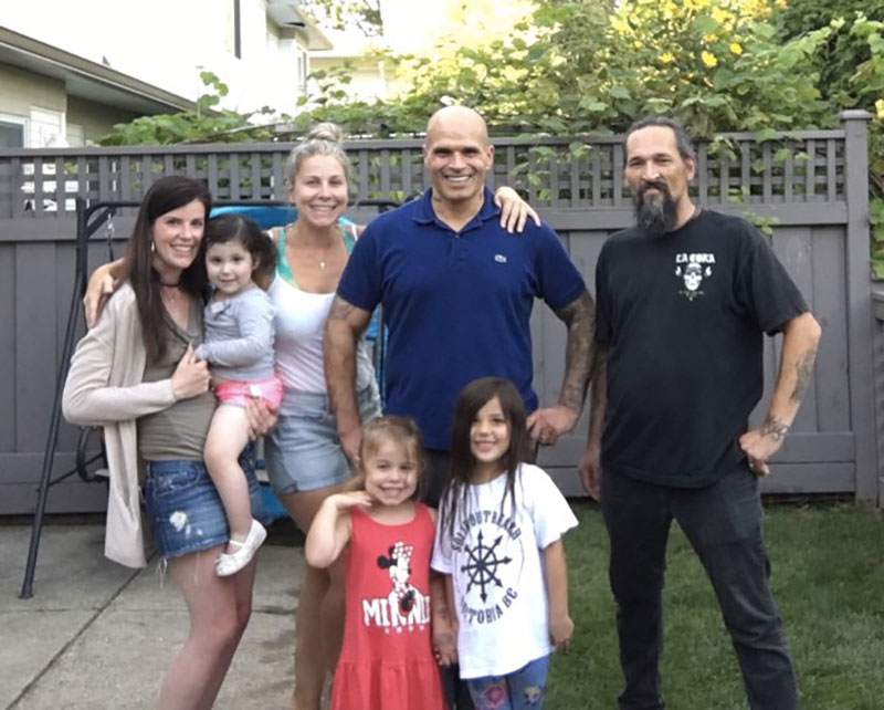 Four adults and three kids pose for a photo in a leafy backyard.