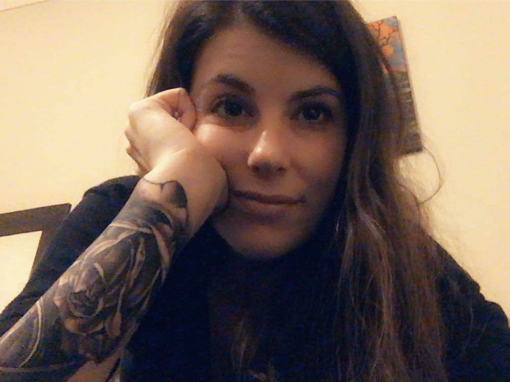 A woman takes a selfie with one hand resting on her cheek. She looks thoughtful and content. She has a large tattoo on her forearm.
