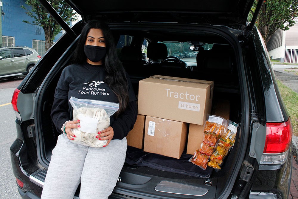 A woman wearing a face mask leans against the back of her vehicle with boxes of food from the Tractor kitchen. She’s holding a bag of cooked chicken which will go on to feed people in her city, likely that night. 