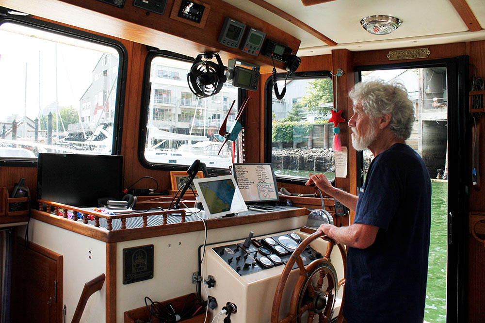 A man with white hair and a beard stands at the helm of his boat as he navigates the waters of False Creek. 