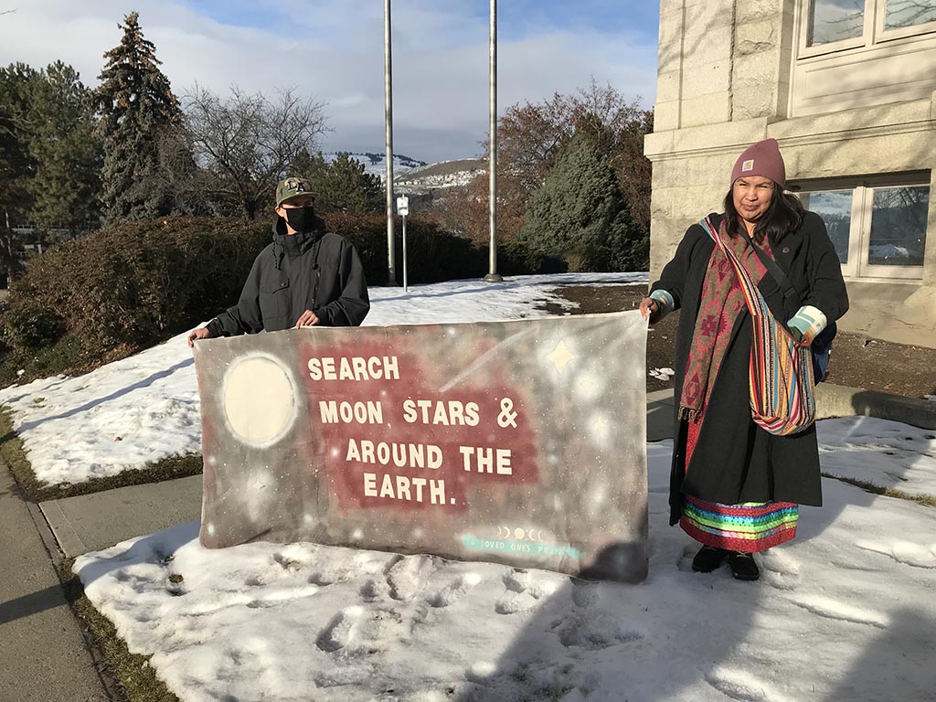 It is snowy. Two people hold a banner reading, ‘Search Moon, Stars & Around the Earth.’