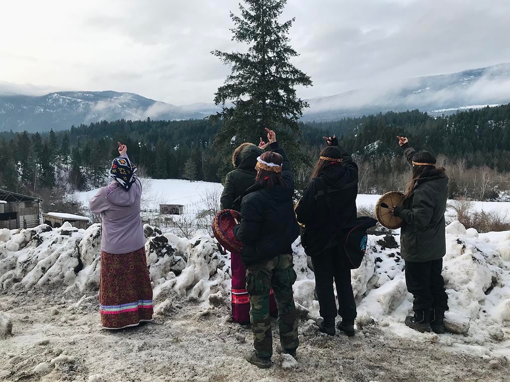On a snowy day, five people stand, backs to the camera looking towards a farm in a valley, holding up their middle fingers.