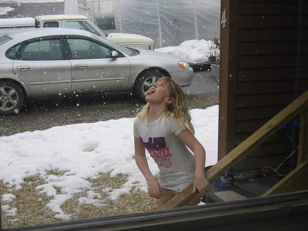 A child in a white T-shirt stands just outside a house during a snowfall, her face turned up towards the sky.