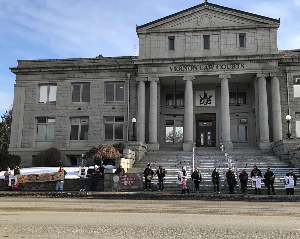 People stand in the snow outside a large grey stone building. The building has lettering reading, Vernon Law Courts.