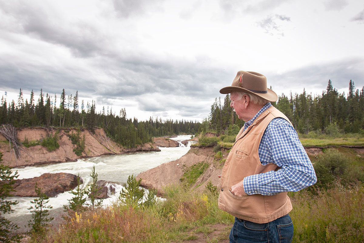 A man wearing a khaki canvas vest and a hat with a feather looks out over a fast-moving river.