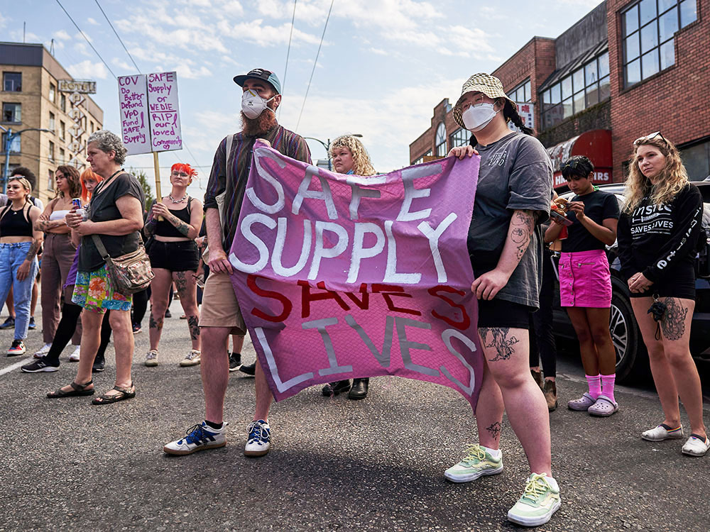 People gather on the street. Many wear face masks. Two people carry a banner saying “Safe Supply Saves Lives.”