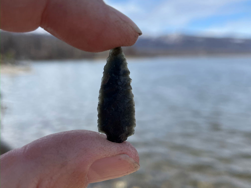 In the foreground, a hand holds a projectile point between thumb and index finger. There is a body of water in the background.