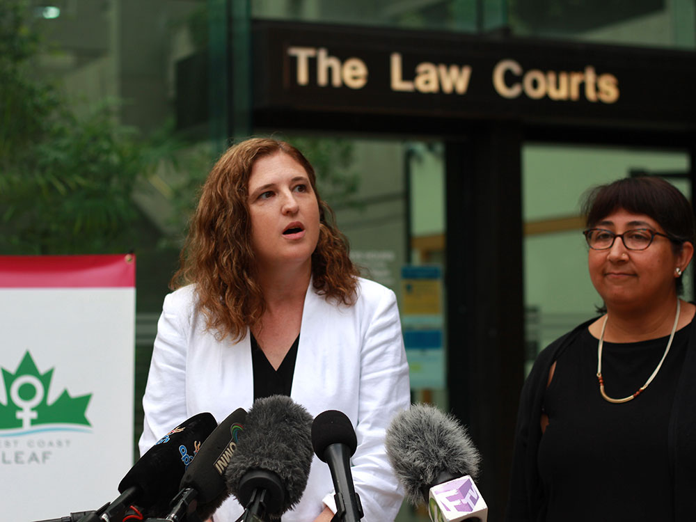 Two women stand outside at a press conference, in front of a microphone. The woman on the left is speaking.
